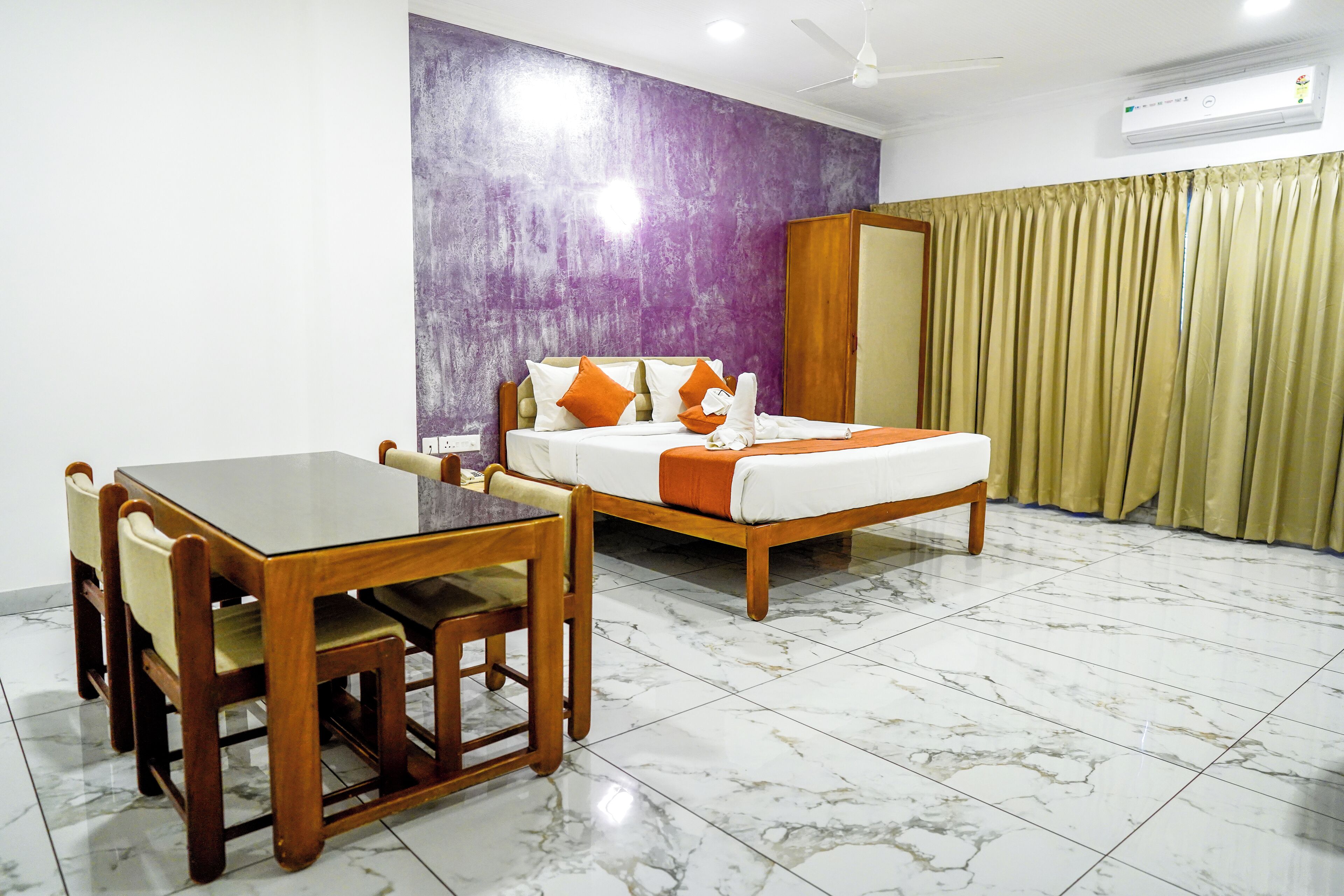 Une chambre d’hôtel avec un lit, une table en bois avec des chaises, un mur d’accent violet et une fenêtre avec des rideaux.