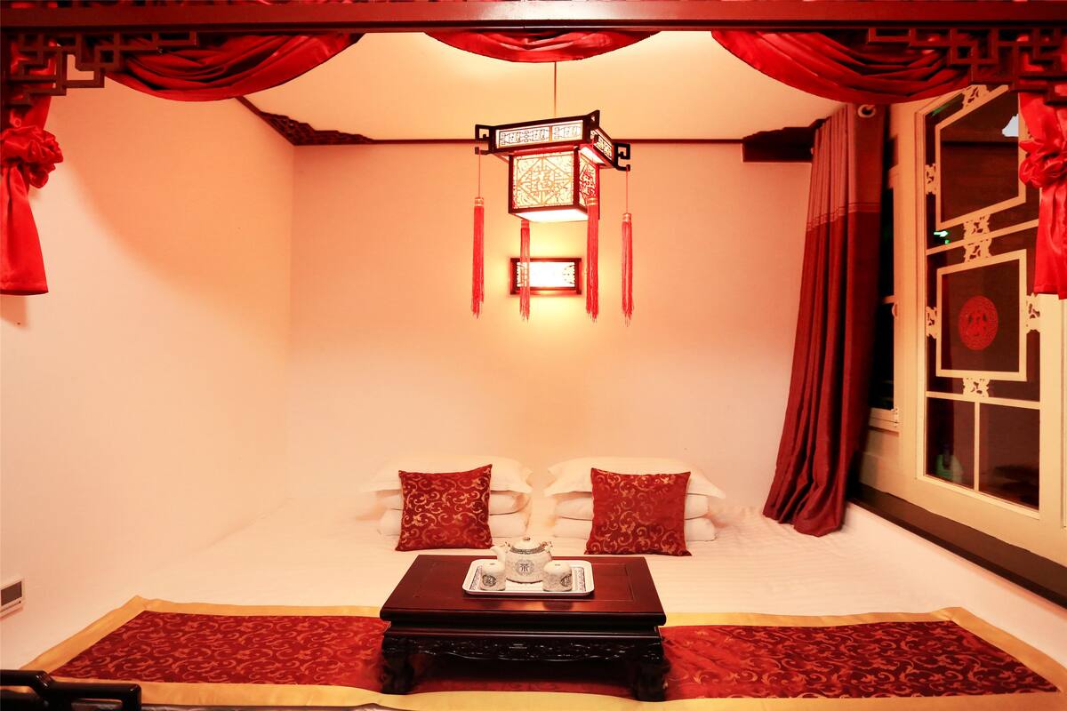 A room with a bed, a bedside table with a tray of tea, red curtains, and a traditional hanging lantern.