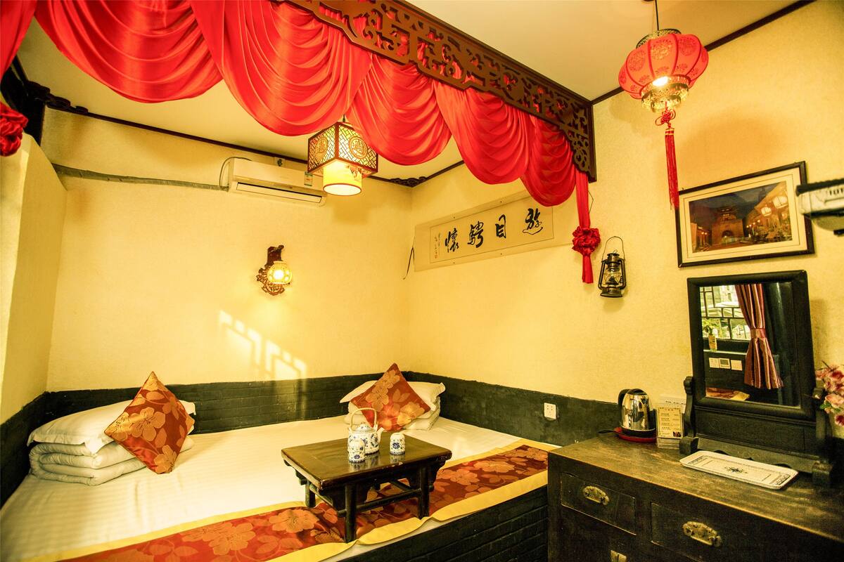 A hotel room with a bed, a small table with teapots, a mirror, and a red lantern.