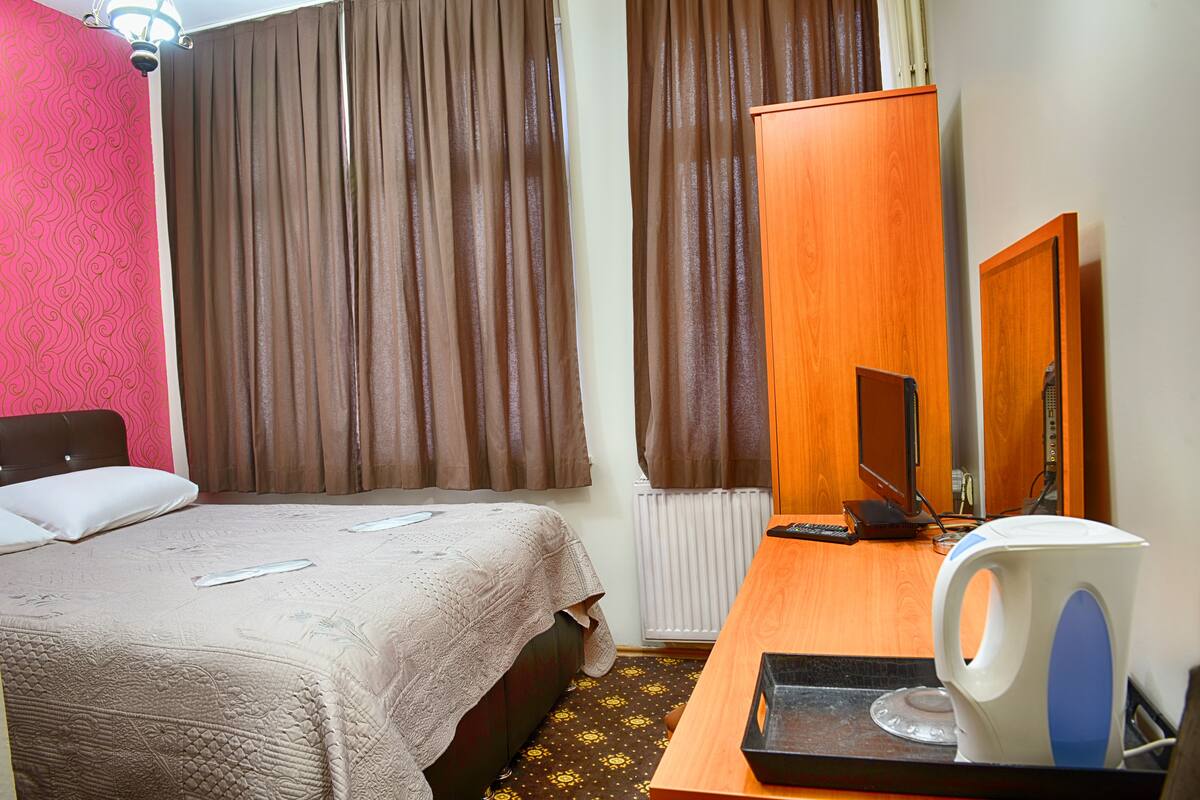 A hotel room with a bed, a desk with a kettle, a mirror, and curtains.