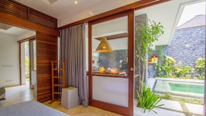 A modern bedroom with a large sliding glass door leading to a pool area, a wooden slatted wall, and a hanging light fixture. - Villa Vishuddha Ajna (Seminyak)