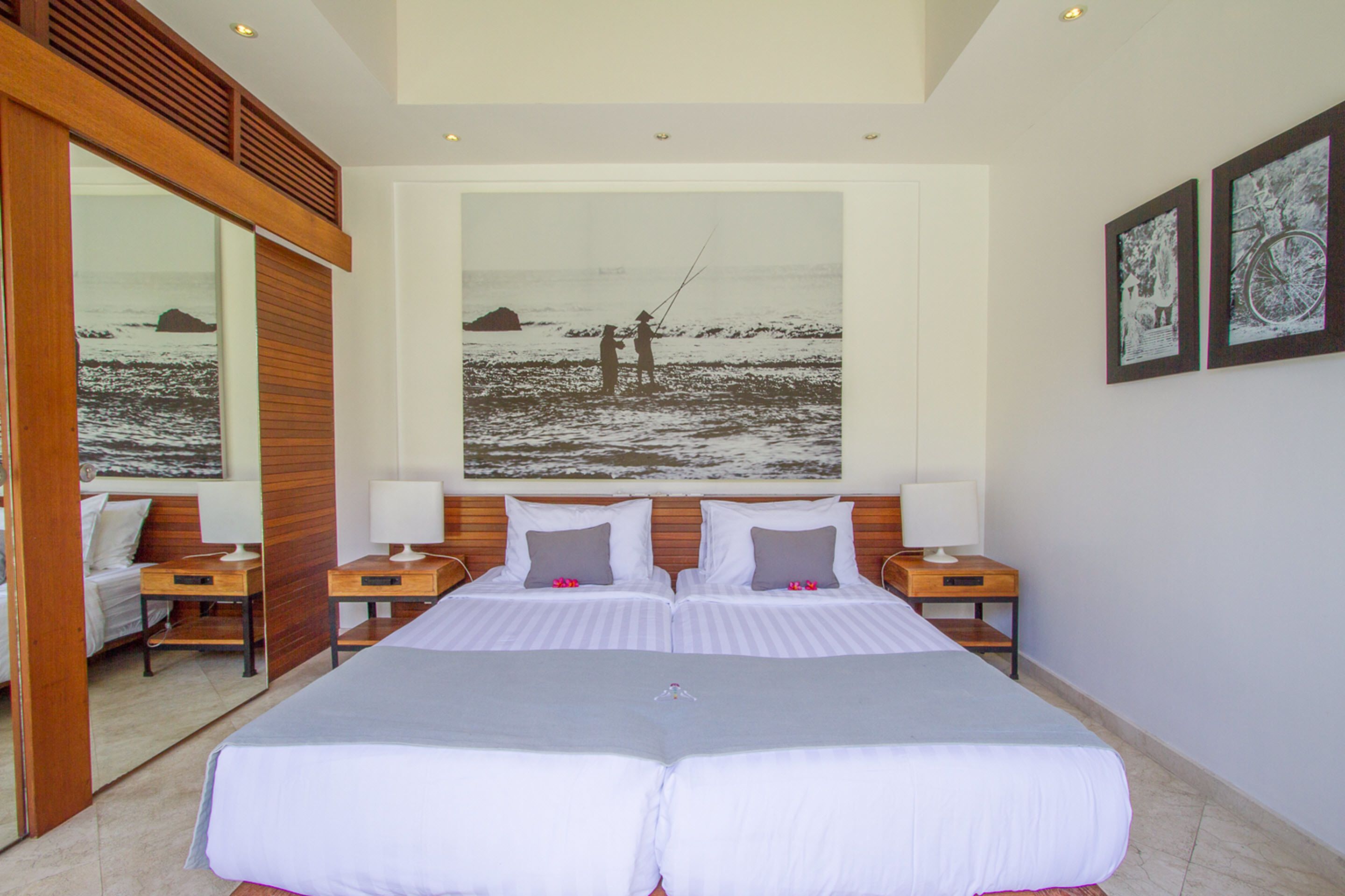 A double bed with a grey bedspread, two bedside tables with lamps, a large framed photograph of a beach scene, and a wooden ceiling.