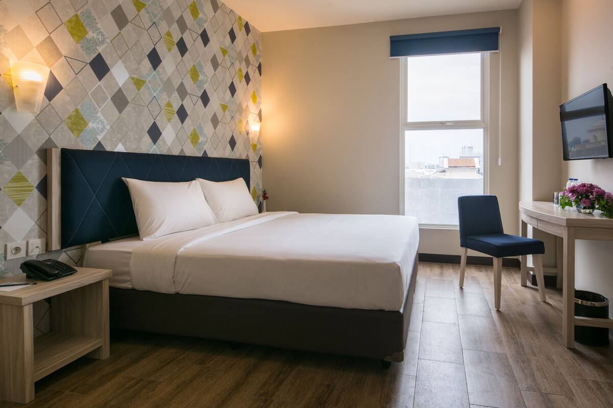 Hotellihuone, jossa on suuri sänky, työpöytä, tuoli, televisio ja ikkuna, josta on näkymä rakennuksiin.