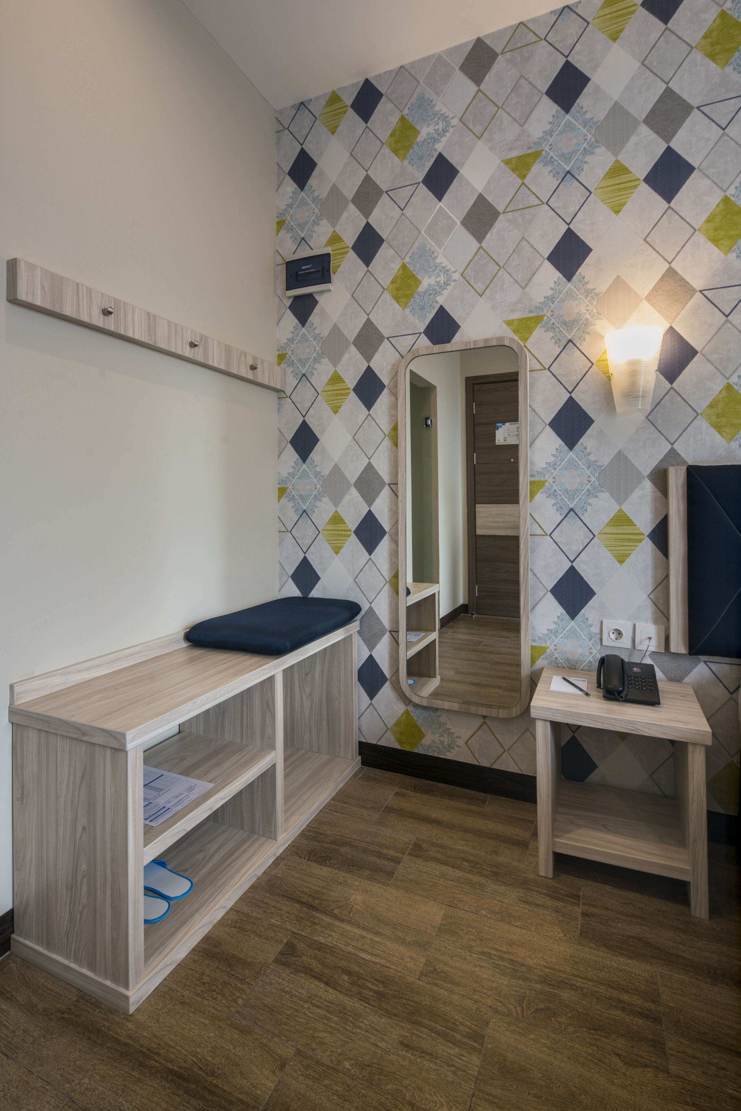 A hotel room with a wooden vanity, a mirror, a shelf with a blue cushion, a small table with a telephone, and a patterned wall.