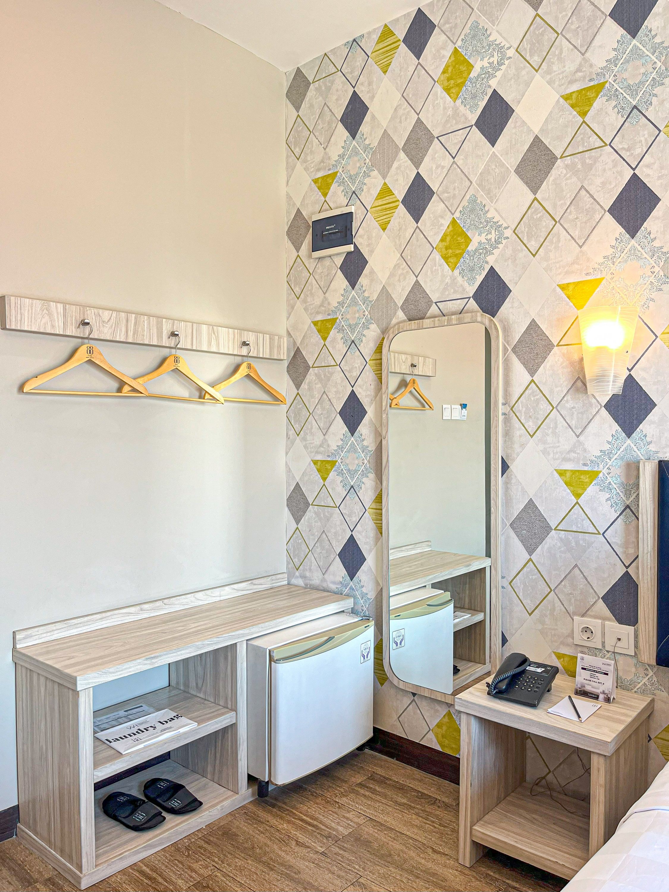 A hotel room with a wooden vanity, a mirror, a small desk with a phone and a sign, and a wall with a geometric patterned wallpaper.
