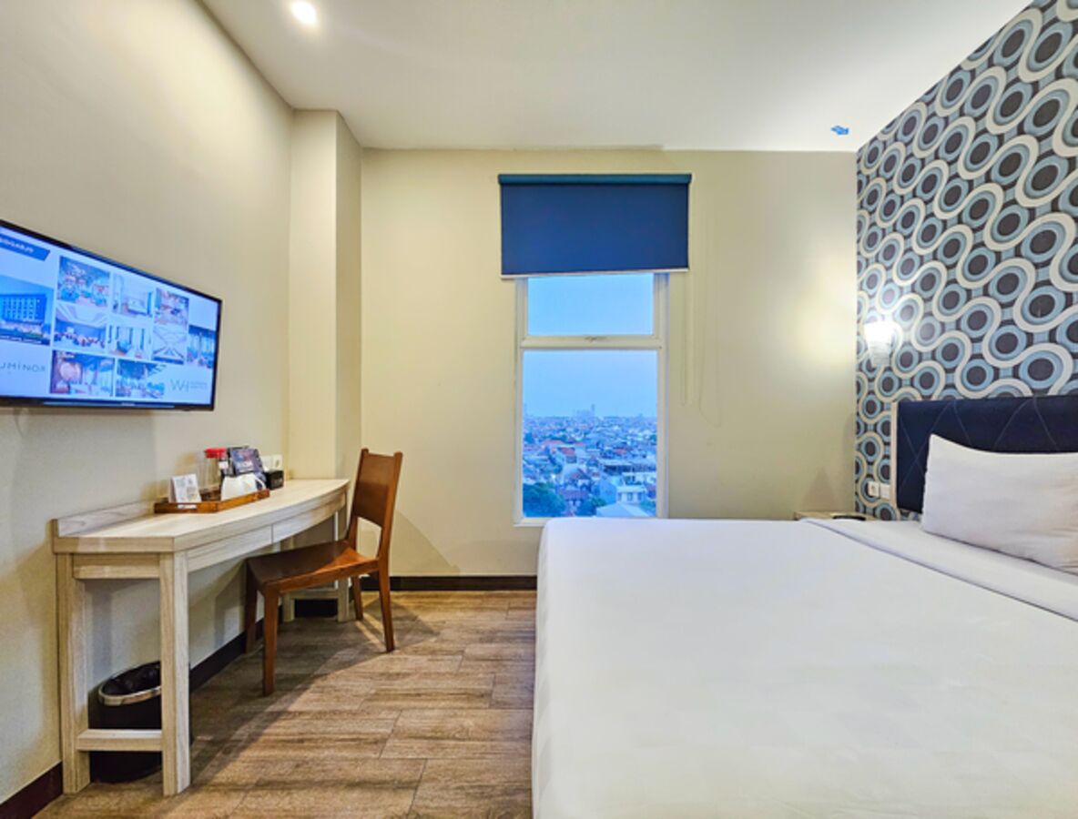 Ein Hotelzimmer mit einem Bett, einem Schreibtisch mit Stuhl, einem an der Wand befestigten Fernseher und einem Fenster mit Blick auf die Stadt.