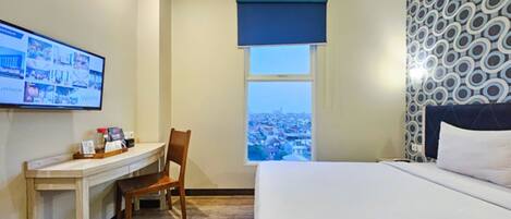 Ein Hotelzimmer mit einem Bett, einem Schreibtisch mit Stuhl, einem an der Wand befestigten Fernseher und einem Fenster mit Blick auf die Stadt.