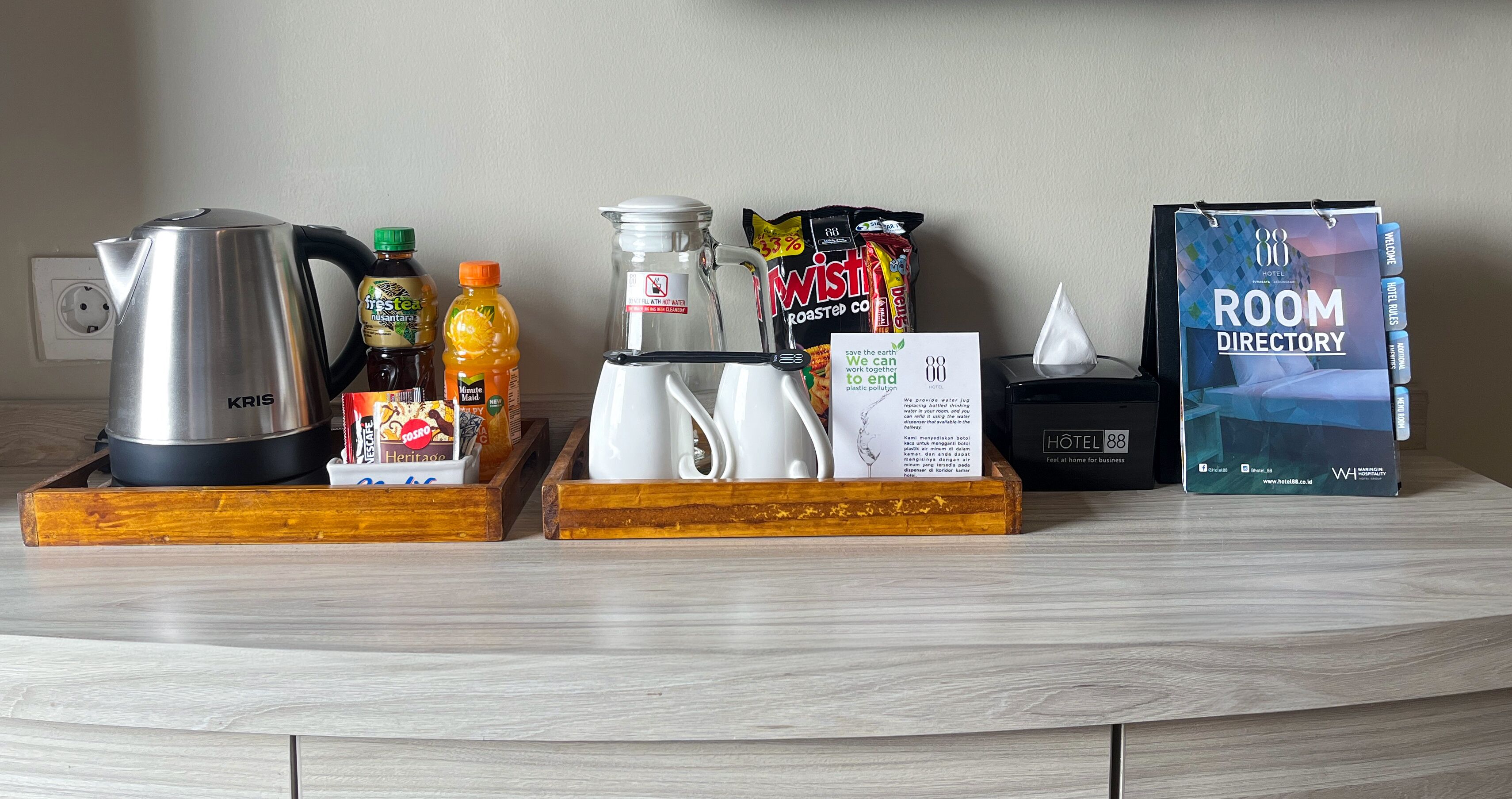 A hotel reception desk with a room directory, a kettle, a coffee maker, a bottle of orange juice, a glass pitcher, a bag of chips, and a tissue box.