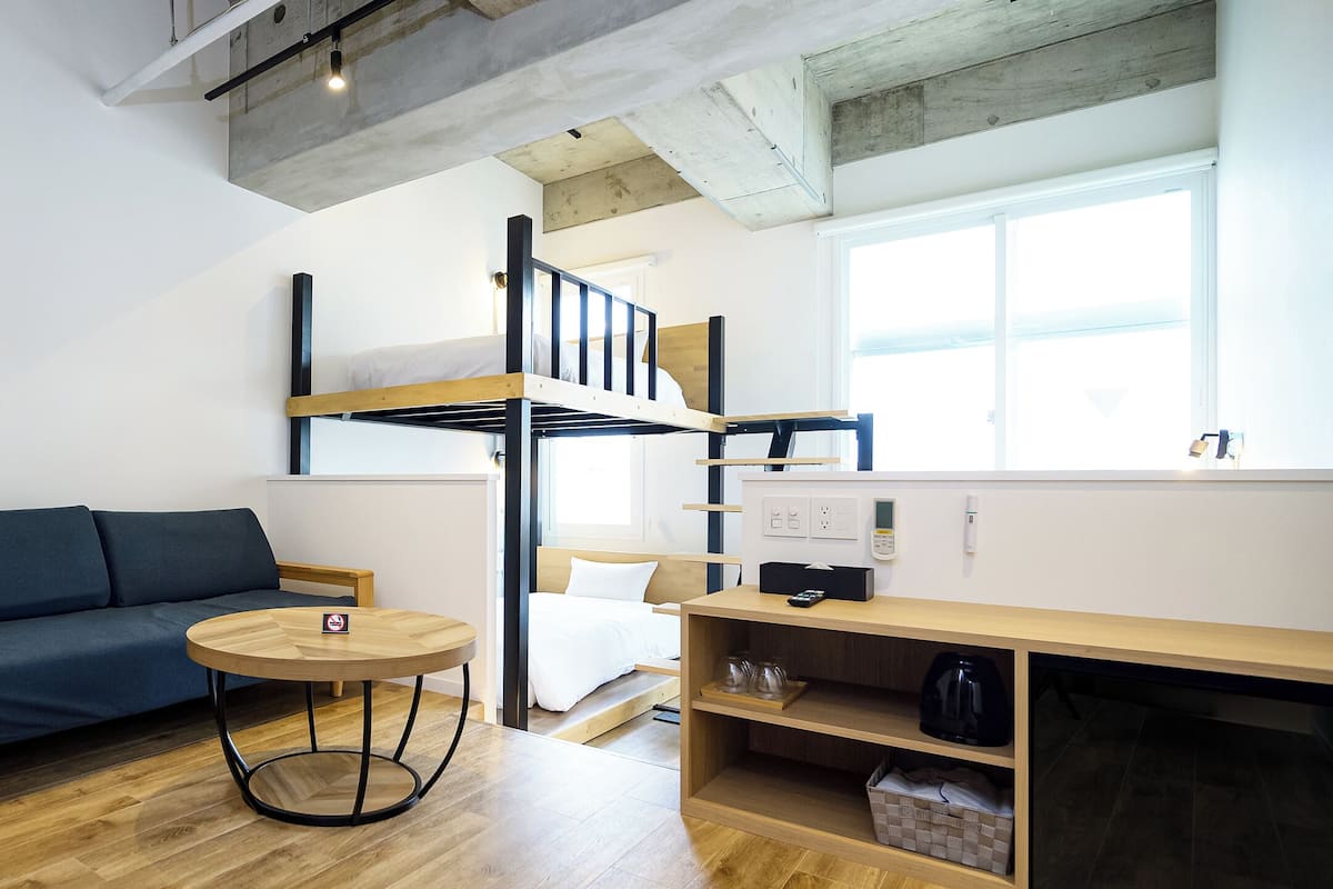 A compact room with a bunk bed, a sofa, a small round table, and a wooden shelf with a kettle.