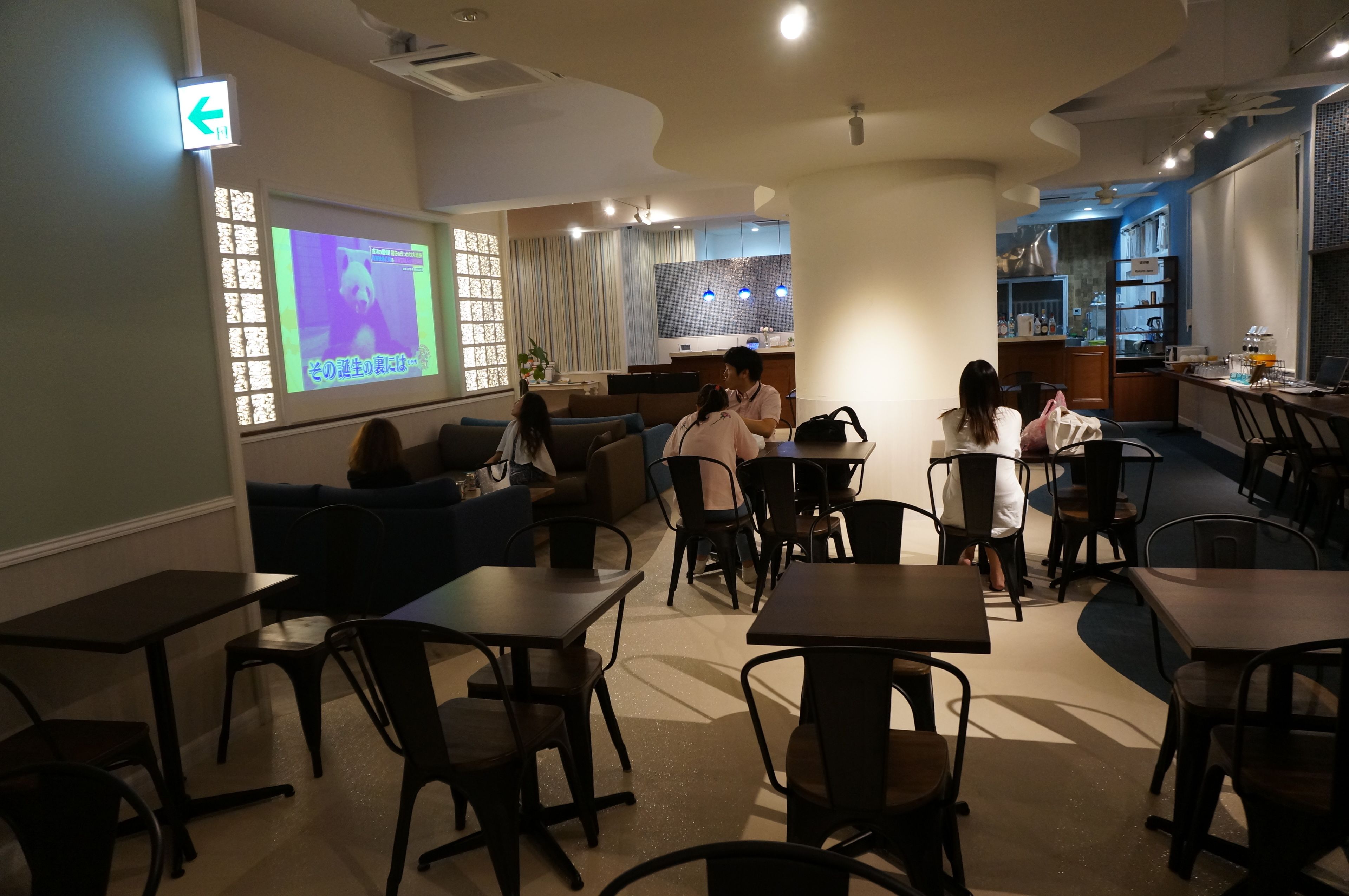 A restaurant with tables and chairs, a kitchen area, and a projection screen displaying a video.