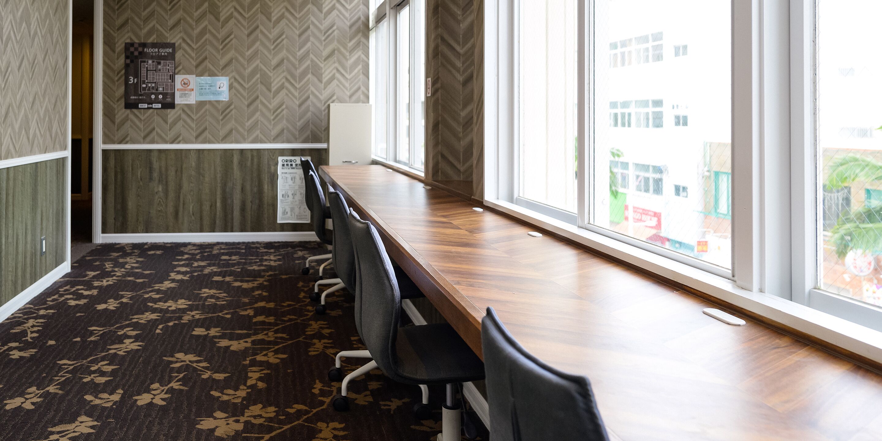 Un salón de conferencias con una larga mesa, sillas, una alfombra estampada con motivos florales y un ventanal con vistas a edificios.