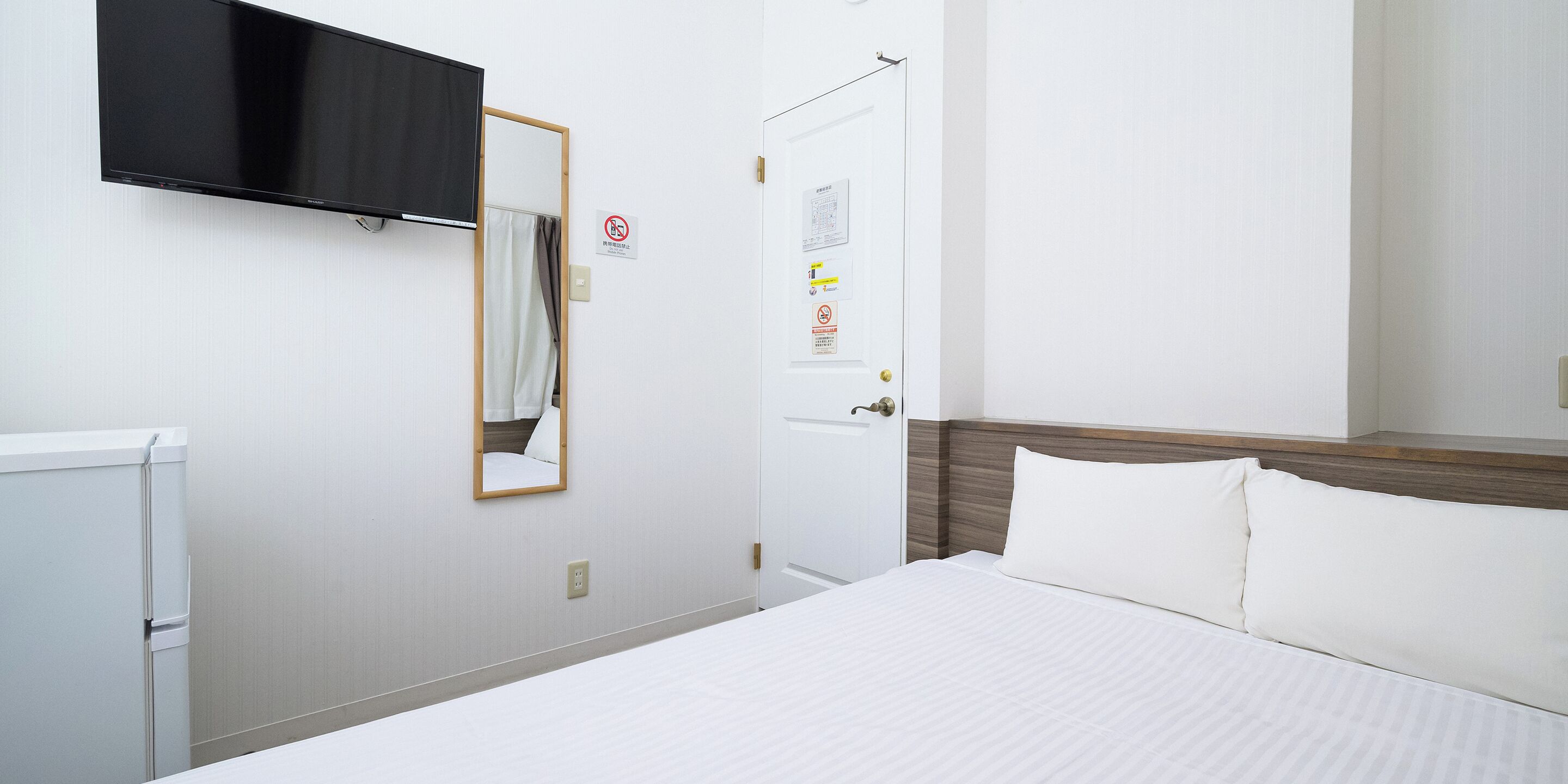 A hotel room with a bed, a flat-screen TV mounted on the wall, a door with a mirror, and a white wall with a no-smoking sign.