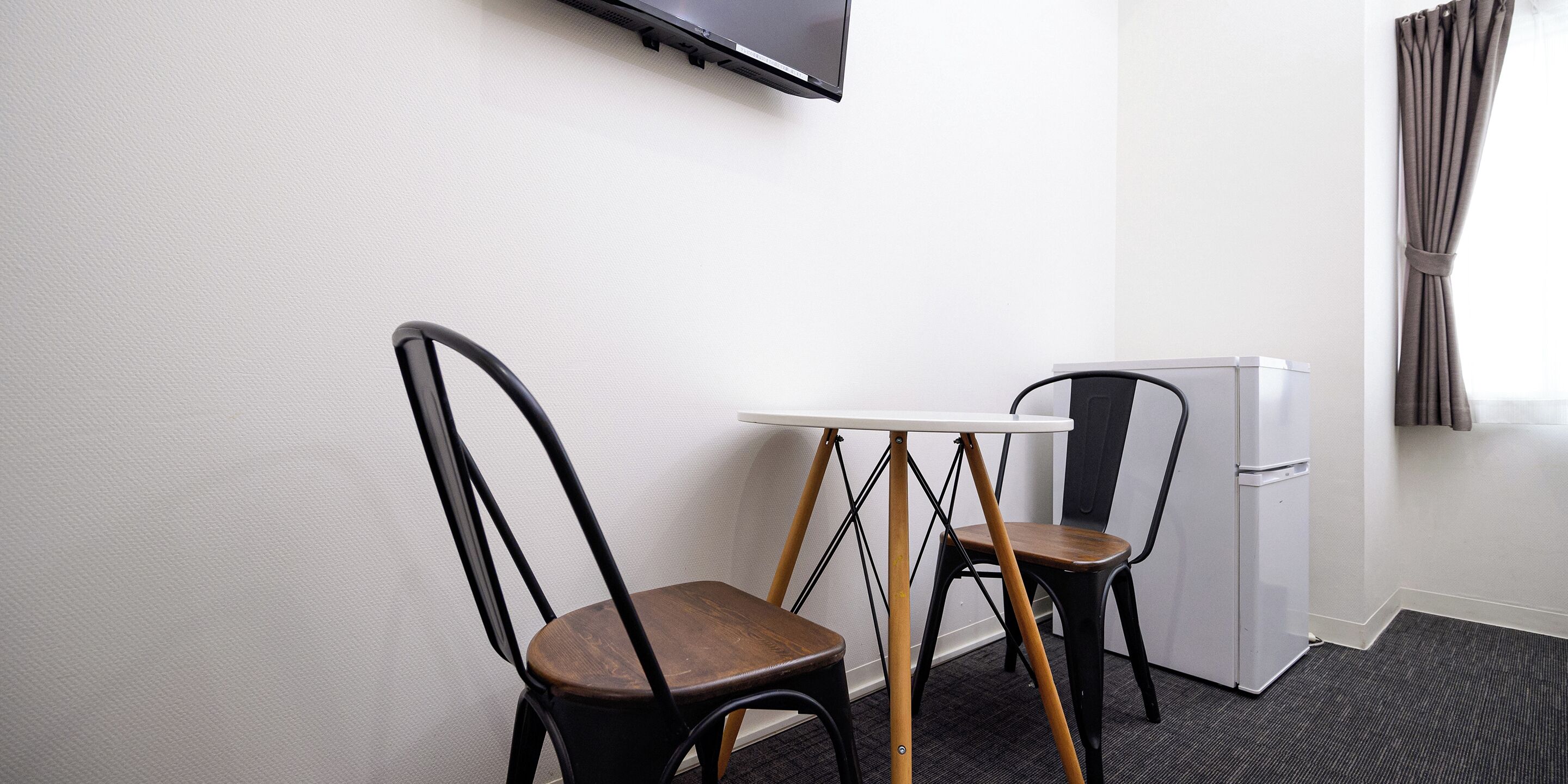 Une petite pièce avec un mur blanc, un téléviseur à écran plat fixé au-dessus d’une table blanche, deux chaises en bois, un réfrigérateur blanc et une fenêtre avec des rideaux.