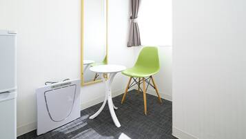 Una habitación pequeña y moderna con un refrigerador blanco, una lavadora compacta, una silla verde, una mesita blanca y un espejo en la pared.