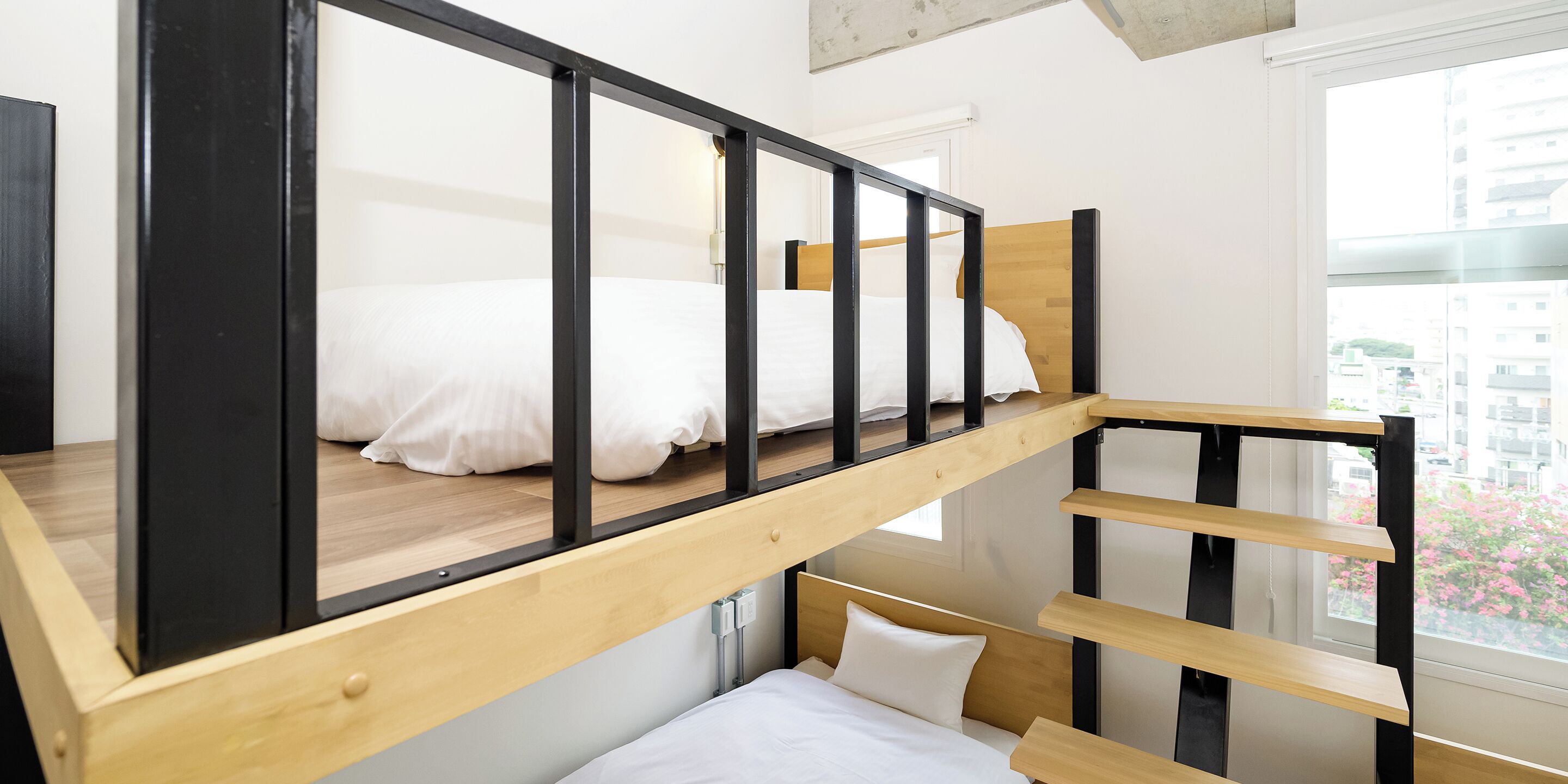 Una cama litera con estructura de madera y ropa de cama blanca, un escritorio de madera pequeño y una ventana con vistas a edificios y flores rosadas.