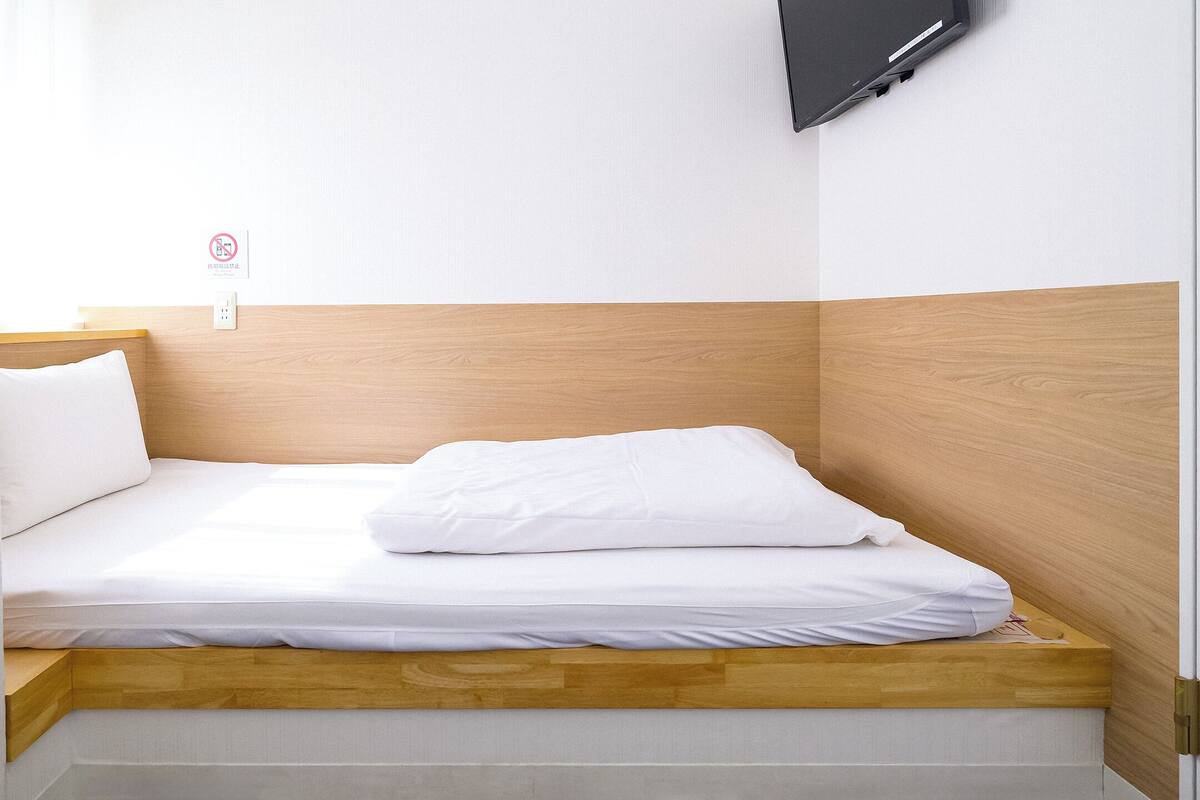 A small, neatly made bed with white bedding in a compact room with a wooden headboard and a mounted TV on the wall.