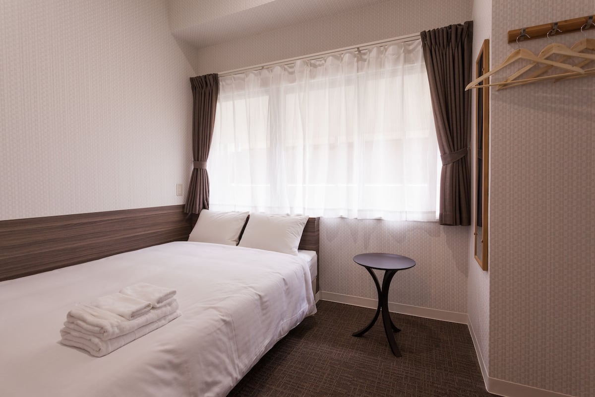 A neatly made bed with white linens and a stack of folded towels, a small round table, and a window with sheer curtains.