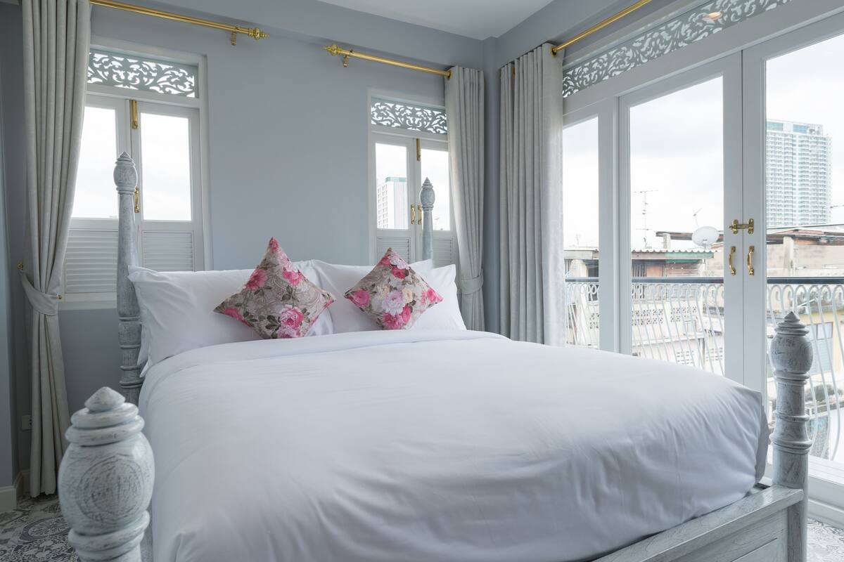 Un lit parfaitement fait, avec du linge de lit blanc et des coussins à motifs floraux, une tête de lit blanche et une vue sur la ville grâce à de grandes fenêtres.