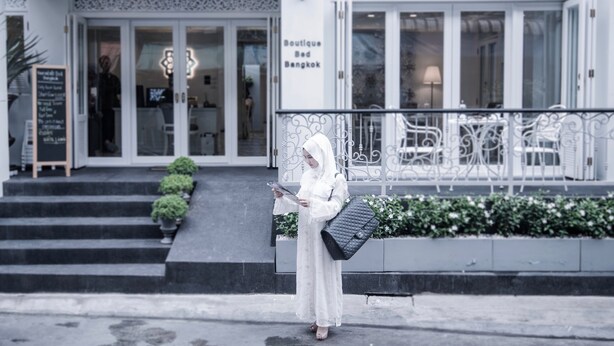 L’entrée d’un hôtel-boutique, avec une enseigne indiquant « Boutique Bed Bangkok », des marches menant à l’entrée et une personne debout sur les marches, un téléphone à la main.