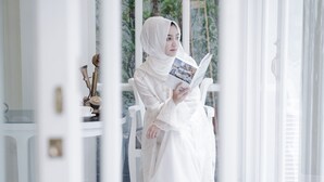 Une personne vêtue d’une robe blanche est assise près d’une fenêtre et lit un livre.