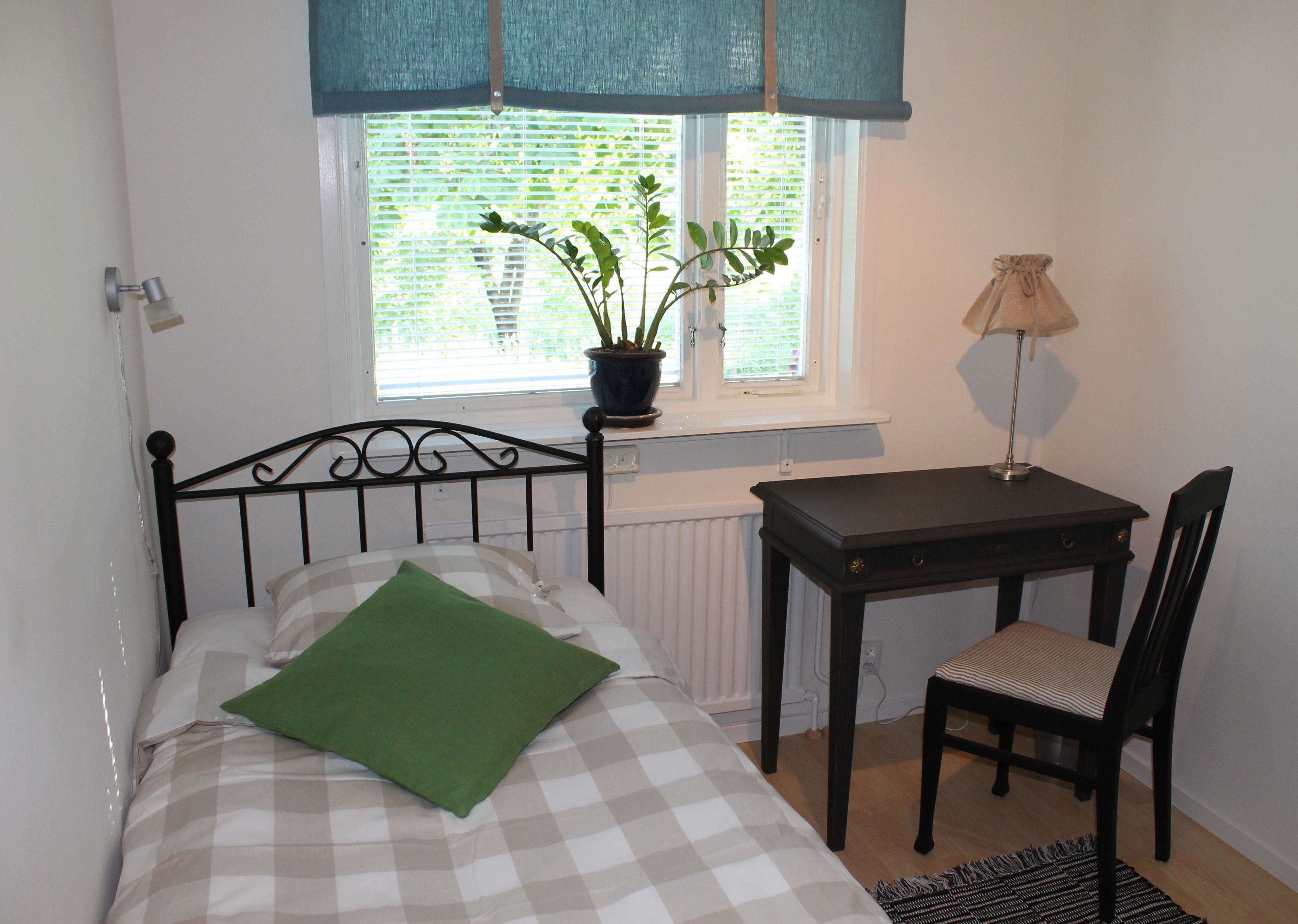 A bedroom with a bed, a desk, a chair, a potted plant, and a window with blinds.