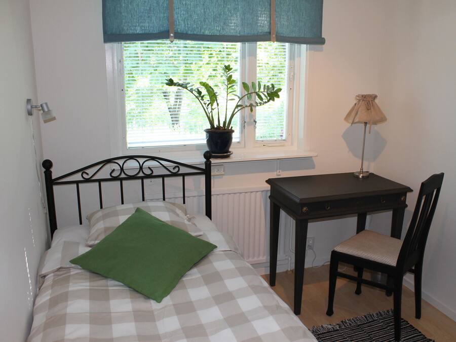 A bedroom with a bed, a desk, a chair, a potted plant, and a window with blinds.