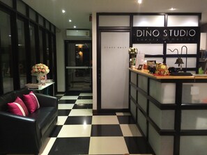 A reception area with a checkered floor, a sofa with pink pillows, and a counter displaying a sign for 'DINO STUDIO Luxury Homestay'. - Dino Studio (Kalasin)