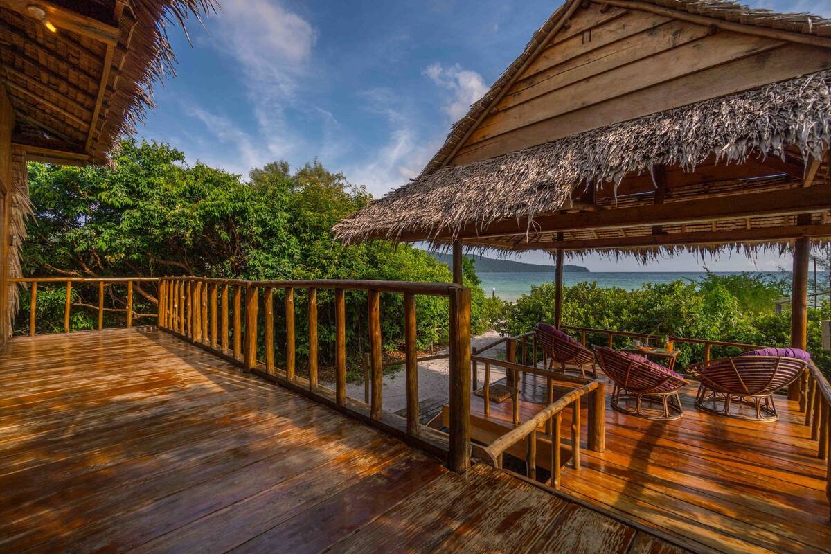 A wooden deck with a thatched roof, overlooking a beach and ocean.
