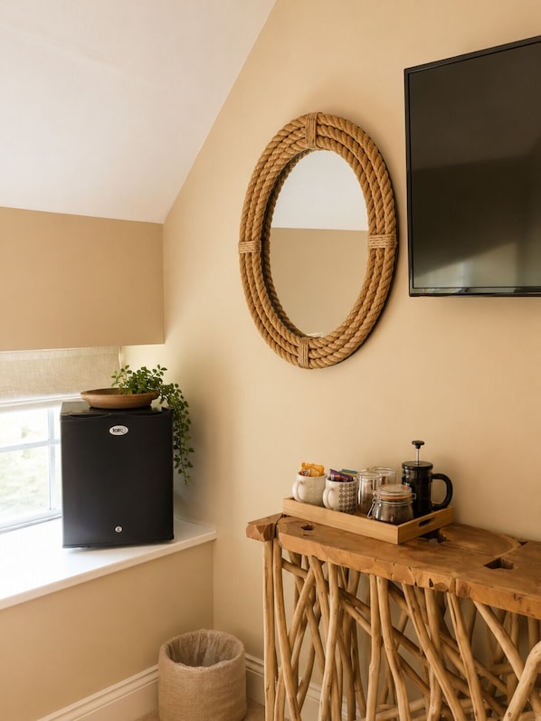 A kitchenette with a black refrigerator, a wooden table with a tray of items, a woven mirror, and a TV on the wall.