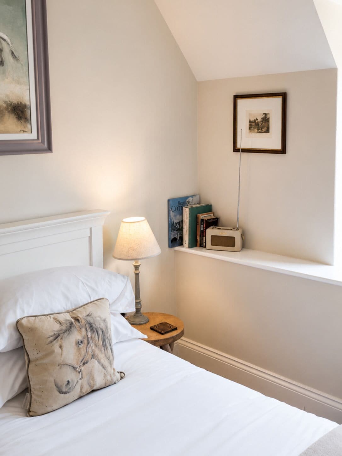 Un dormitorio con cama, almohadas, una lámpara de noche, una estantería con libros y un cuadro enmarcado, y una mesita con un radio.