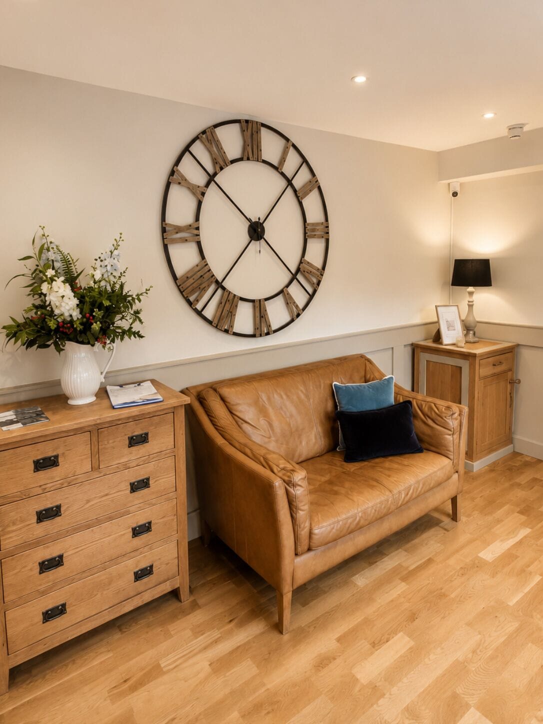 A room with a large wall clock, a wooden cabinet with drawers, a leather sofa, and a small table with a lamp.