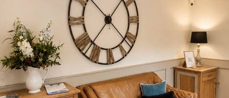 A room with a large wall clock, a wooden cabinet with drawers, a leather sofa, and a small table with a lamp.
