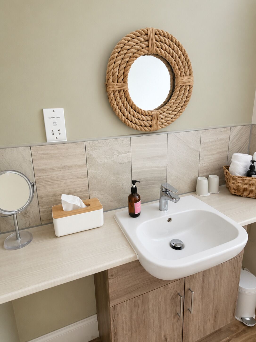 A bathroom with a round woven mirror, a white sink, a soap dispenser, a tissue box, and a trash bin.