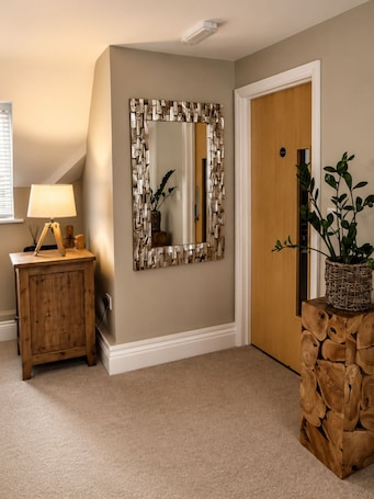 A hallway with a wooden cabinet, a lamp, a mirror with a geometric frame, a door, and a plant in a woven basket.