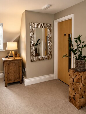 A hallway with a wooden cabinet, a lamp, a mirror with a geometric frame, a door, and a plant in a woven basket.
