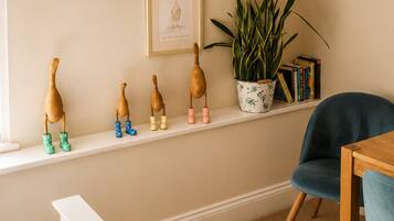 A room with a white bench, a blue chair, a wooden table, a potted plant, and a shelf with wooden figures and books.