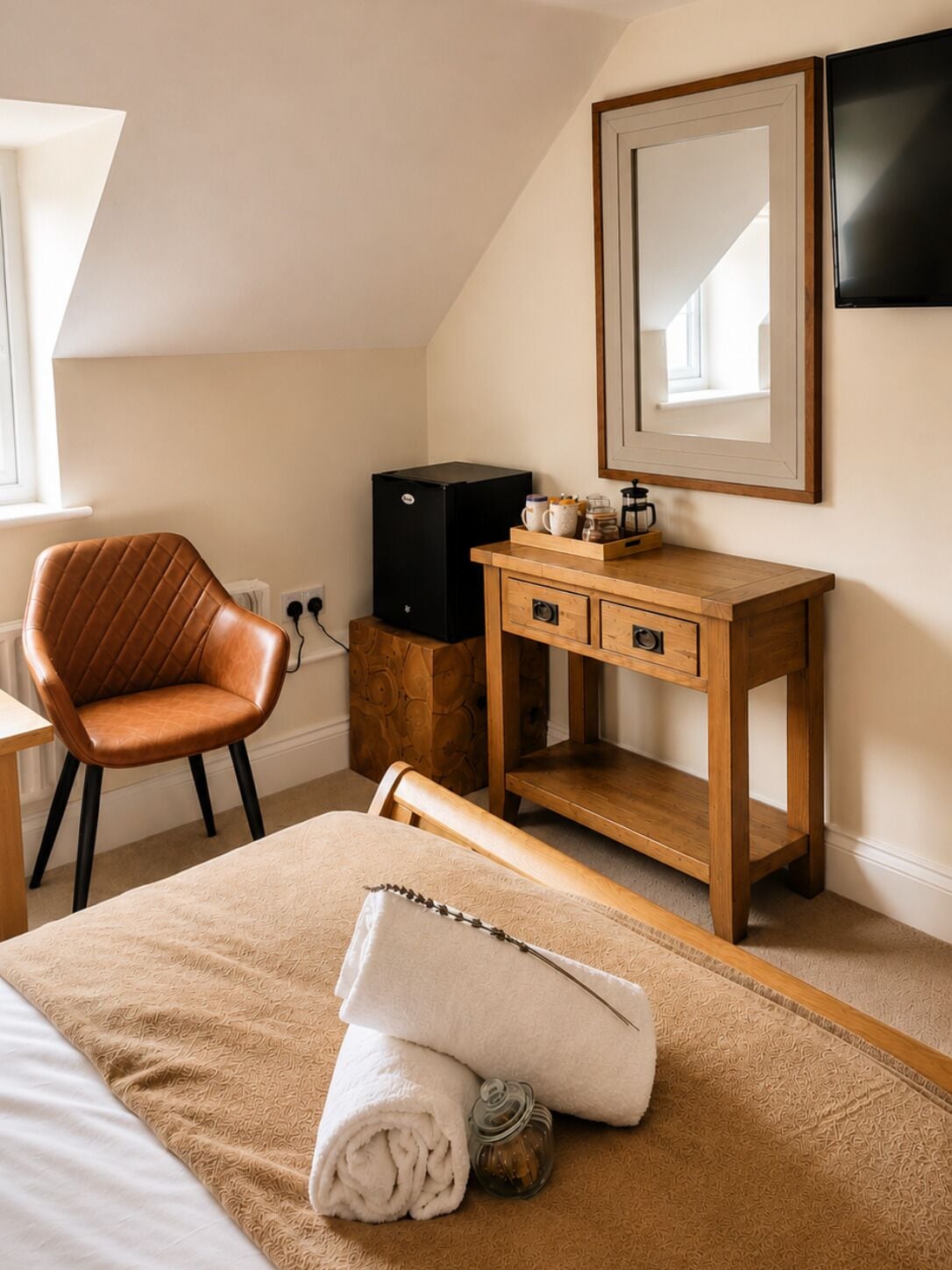 A bedroom with a bed, a wooden bedside table, a chair, a black refrigerator, and a mirror on the wall.