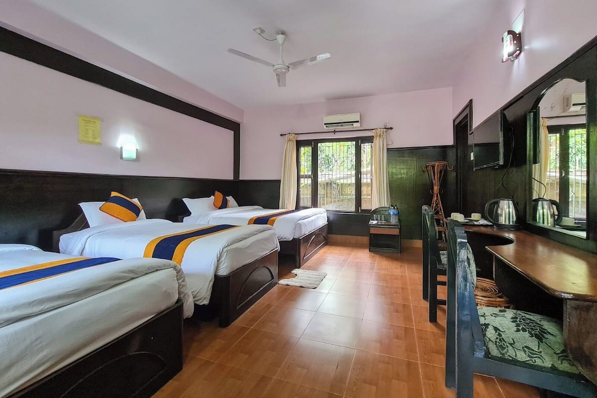 A hotel room with three beds, a ceiling fan, an air conditioning unit, a small table with a kettle, and a large mirror.