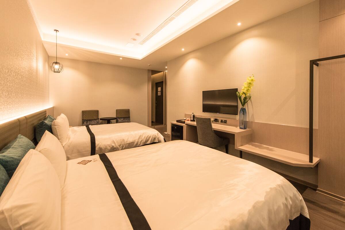 A hotel room with two beds, a desk, a chair, a TV, and a vase with flowers.