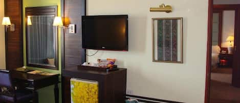 A hotel room with a flat-screen TV mounted on the wall, a small refrigerator, a desk with a chair, and a bedside table with a lamp.