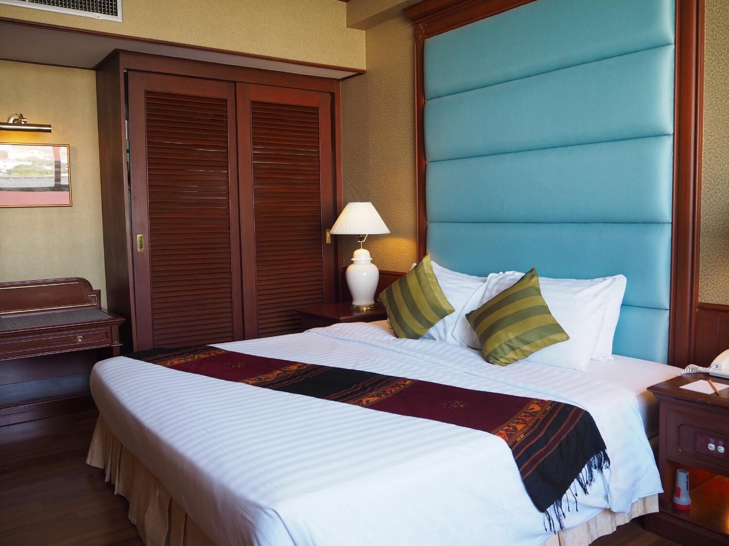 A hotel room with a large bed, a wooden wardrobe, a nightstand with a lamp, and a desk with a telephone.