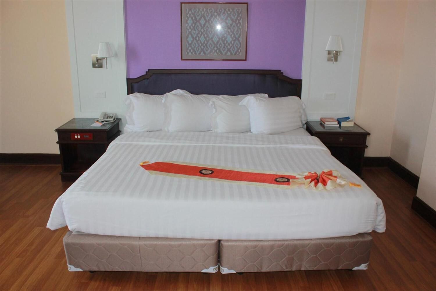 A hotel room with a large bed, two bedside tables, a nightstand, a wall-mounted lamp, and a framed artwork above the bed.