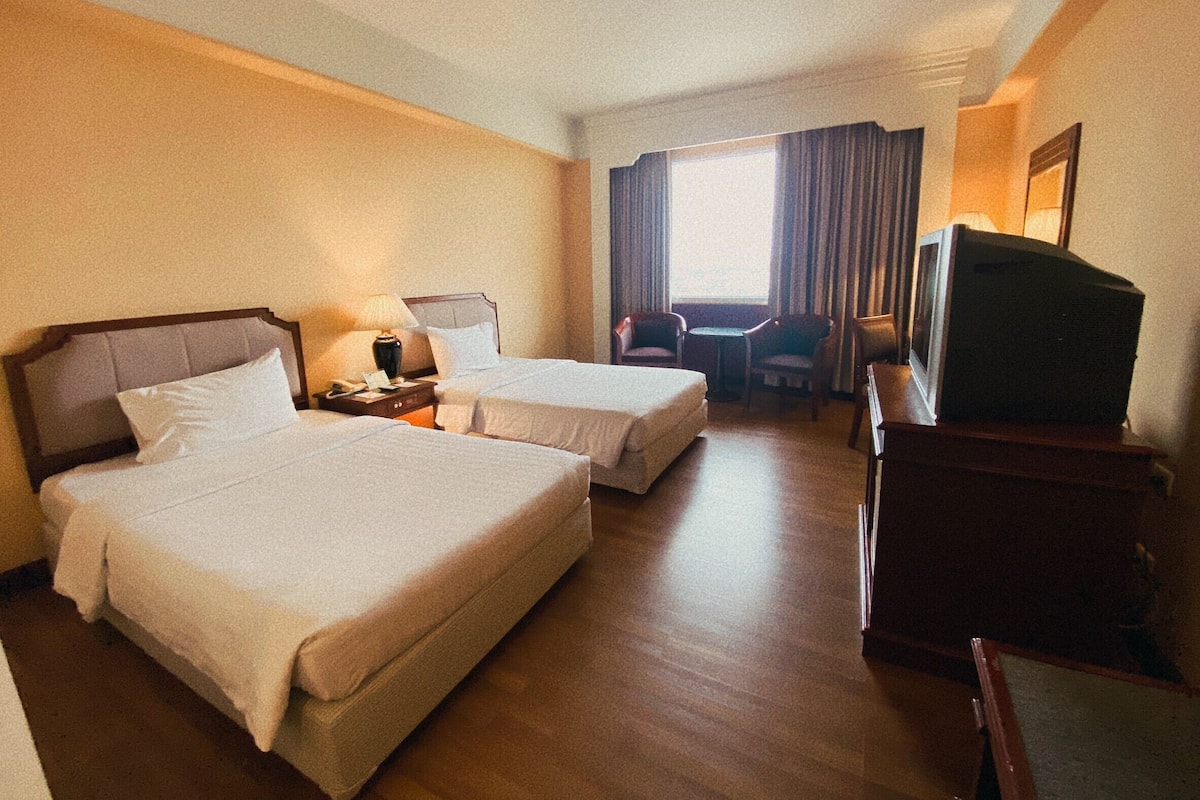 Quarto de hotel com duas camas, uma mesa, uma cadeira, televisão e janela com cortinas.