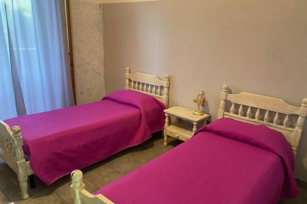 A room with two single beds, each with a wooden headboard and a purple bedspread. There is a small bedside table with a lamp on one side.
