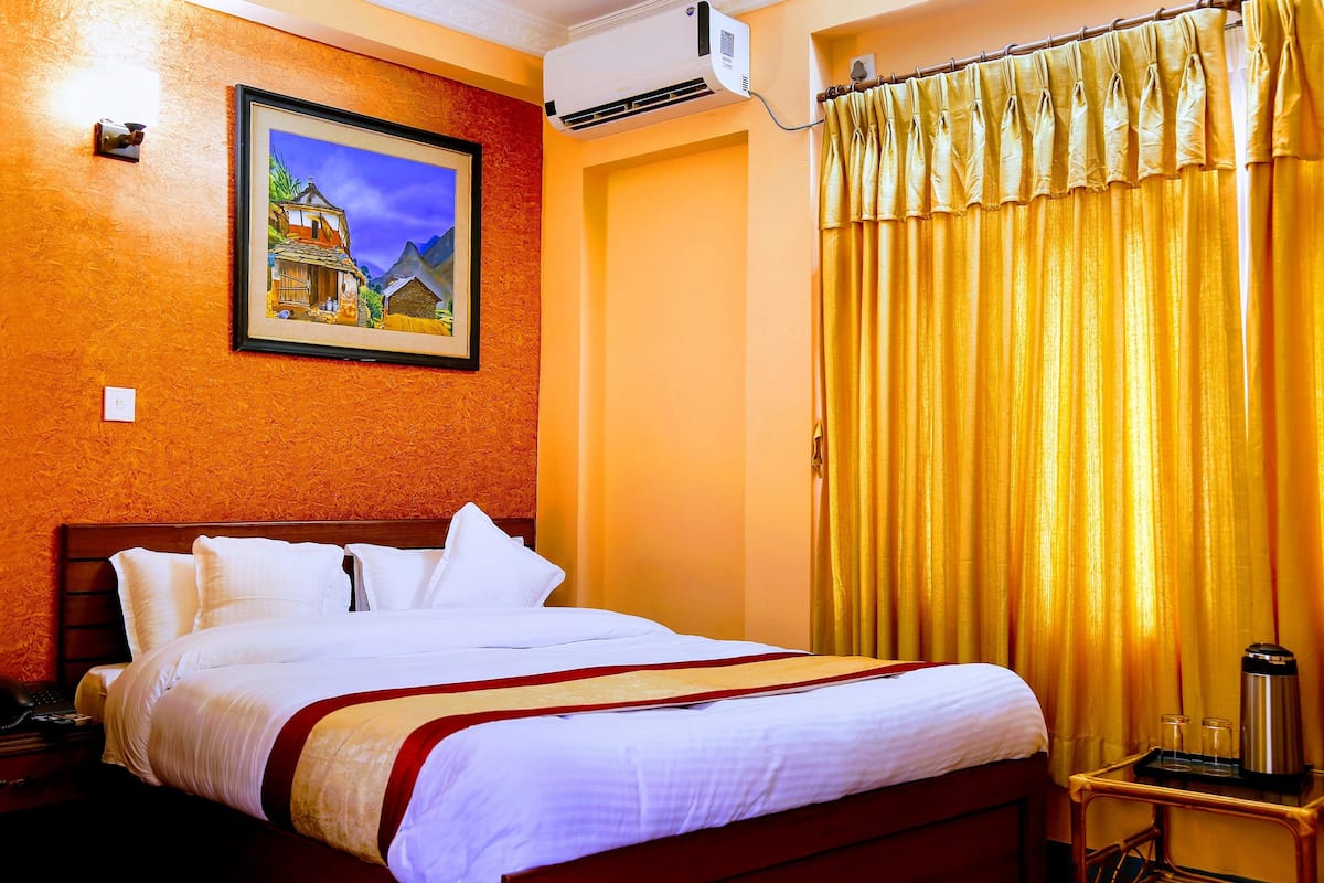 A hotel room with a bed, a painting on the wall, an air conditioning unit, and a curtain.