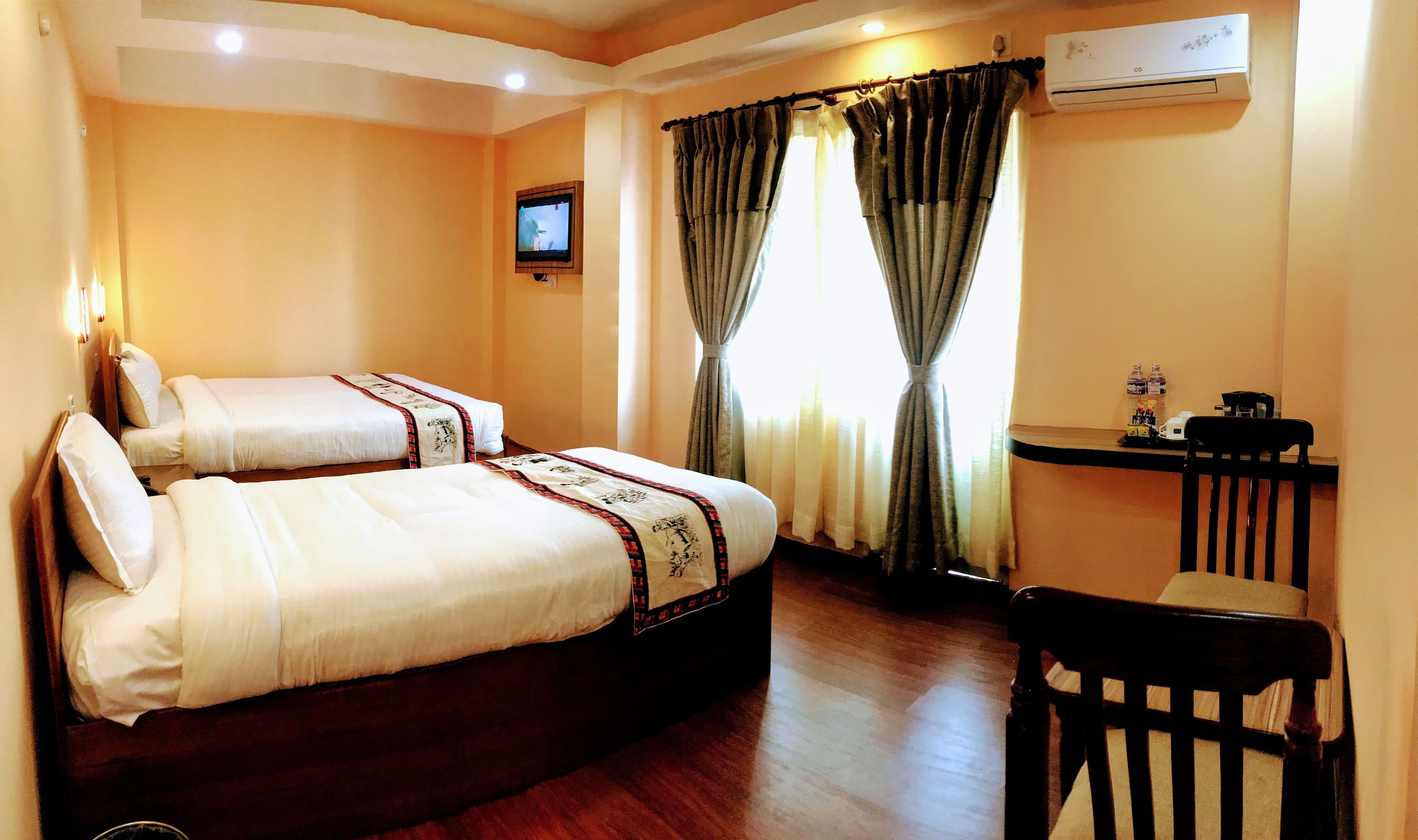 A hotel room with two beds, a desk with a chair, an air conditioner, and a television on the wall.