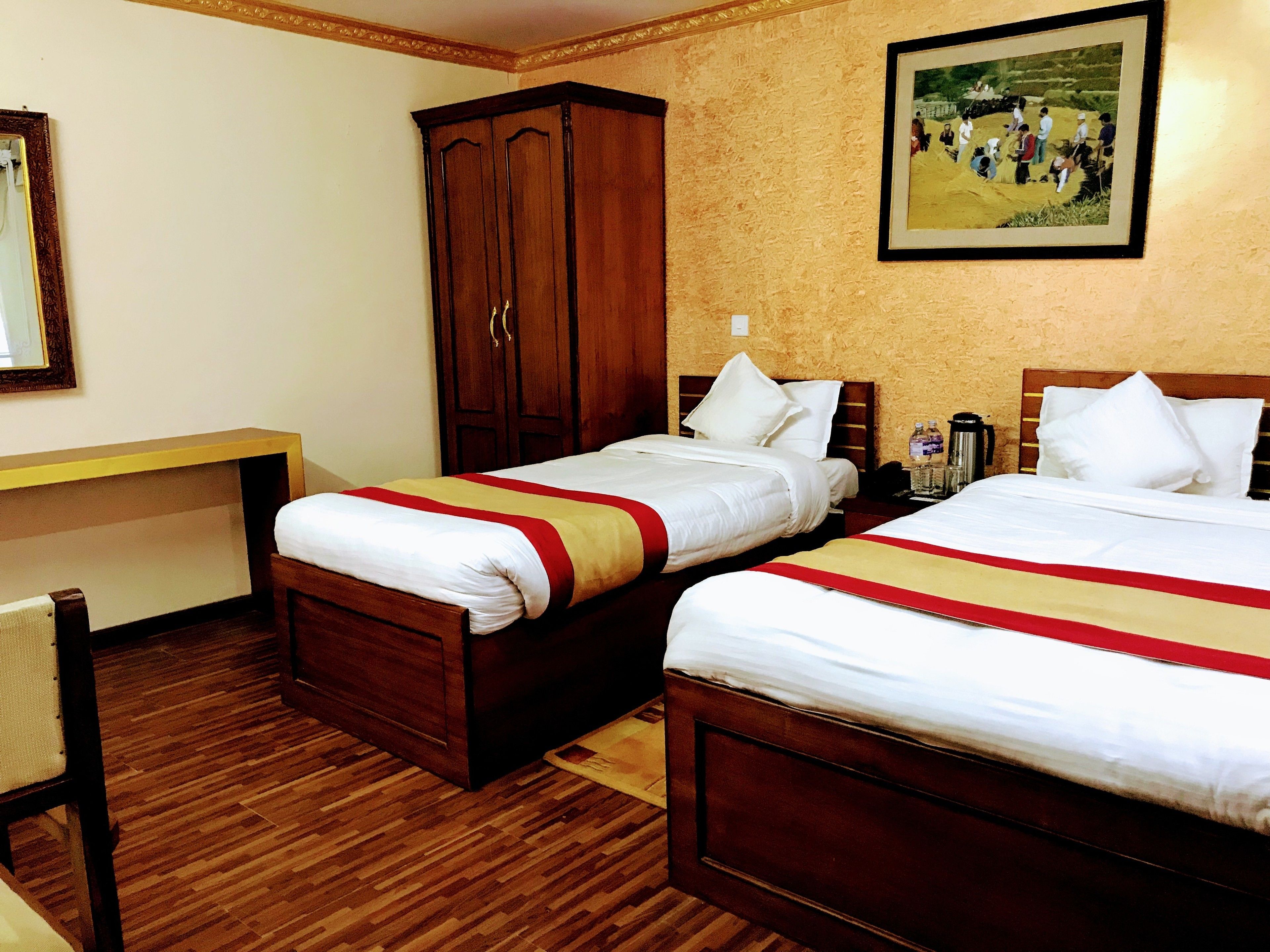 A hotel room with two beds, a wooden wardrobe, a desk, and a framed picture on the wall.