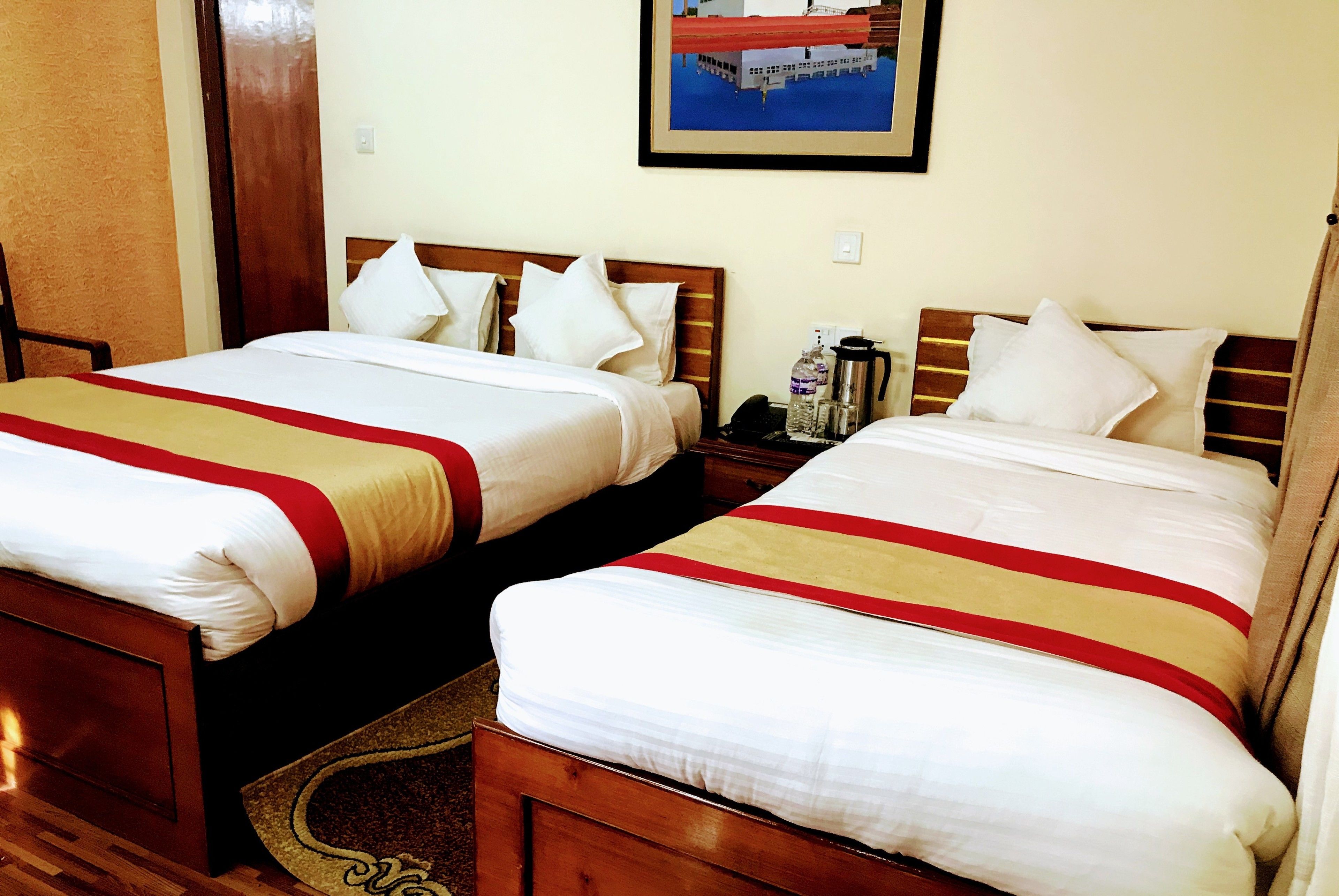 Habitación de hotel con dos camas, cada una con ropa de cama blanca y franjas rojas y beige. Hay una mesita de noche con un teléfono y una botella de agua. Un cuadro enmarcado cuelga de la pared sobre las camas.