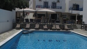 A swimming pool with lounge chairs and umbrellas, surrounded by a building with balconies. - Irene Village (Hersonissos)