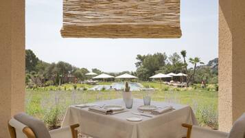 An outdoor dining area with a table set for two, surrounded by chairs, and overlooking a pool and garden.